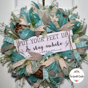 Summer wreath with beachy colors of teal sand and white ribbons, a white wooden sign that reads Put your feet up and stay awhile, with tree ball ornaments that remind you of driftwood, it has a large bow in matching colors, accented by dainty white flowers with green leaves