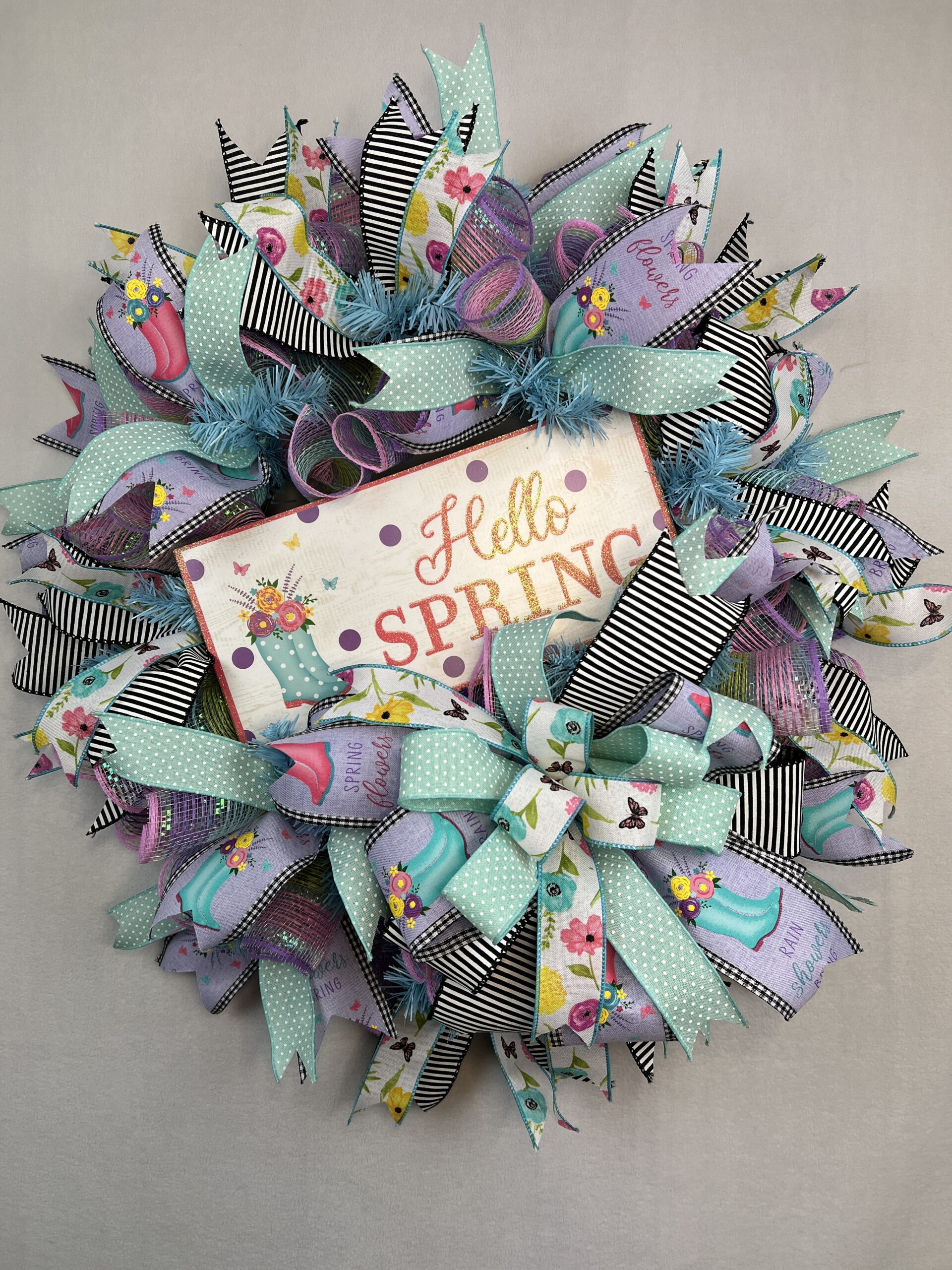 picture of a spring deco mesh wreath with hello spring sign and beautiful spring colored ribbons of purple, teal and whit with flowers