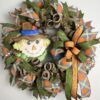 picture of a deco mesh fall scarecrow wreath, with a cute scarecrow face sign and ribbons of orange, blue and yellow plaid ribbon, moss green ribbon, on a base of brown metallic deco mesh. a beautiful bow accents the scarecrow sign and colorful ribbons.