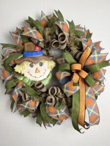 picture of a deco mesh fall scarecrow wreath, with a cute scarecrow face sign and ribbons of orange, blue and yellow plaid ribbon, moss green ribbon, on a base of brown metallic deco mesh. a beautiful bow accents the scarecrow sign and colorful ribbons.