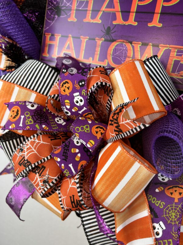 Halloween wreath with a Happy Halloween sign, halloween ribbons of orange stripes, black and white stripes, purple ribbon with eyeballs and skull/candy corn/bats and spider web ribbon. A large bow made from the same ribbon adorns the wreath.