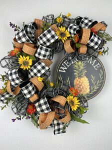 pineapple welcome grapevine wreath, welcome sign with a beautiful pineapple on it, buffalo plaid ribbons and dark gold ribbons wind through the wreath, sunflowers and with smaller burnt red flowers adorn the wreath, adding some greens rounds out this gorgeous wreat
