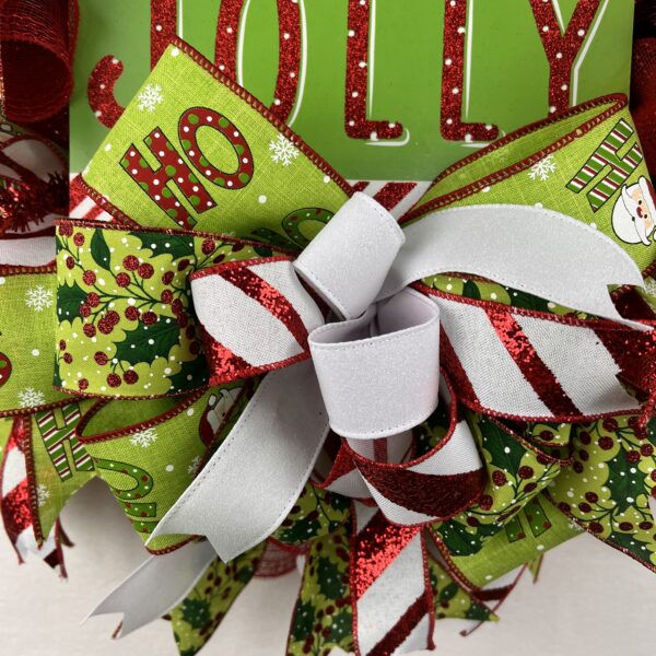 Christmas holiday wreath with red and green Holly Jolly sign in the center of the wreath, with santa ho ho ho ribbons, mistle toe ribbons, red and white stripped ribbon, white ribbon, a bow of matching ribbons is under the sign, red deco mesh is the base for this wreath